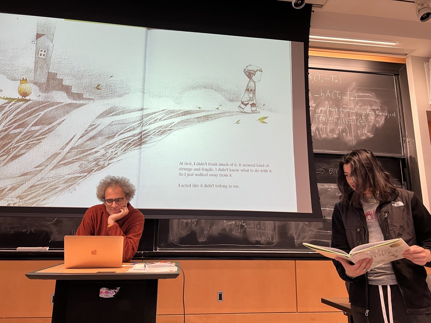 A student reads a picture book aloud in an MIT lecture hall, its illustration projected on the screen while Graham M. Jones listens