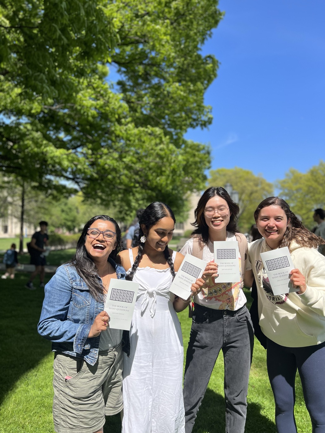 Four students standing together on a sunlit lawn, laughing, each holding a copy of the book their class made